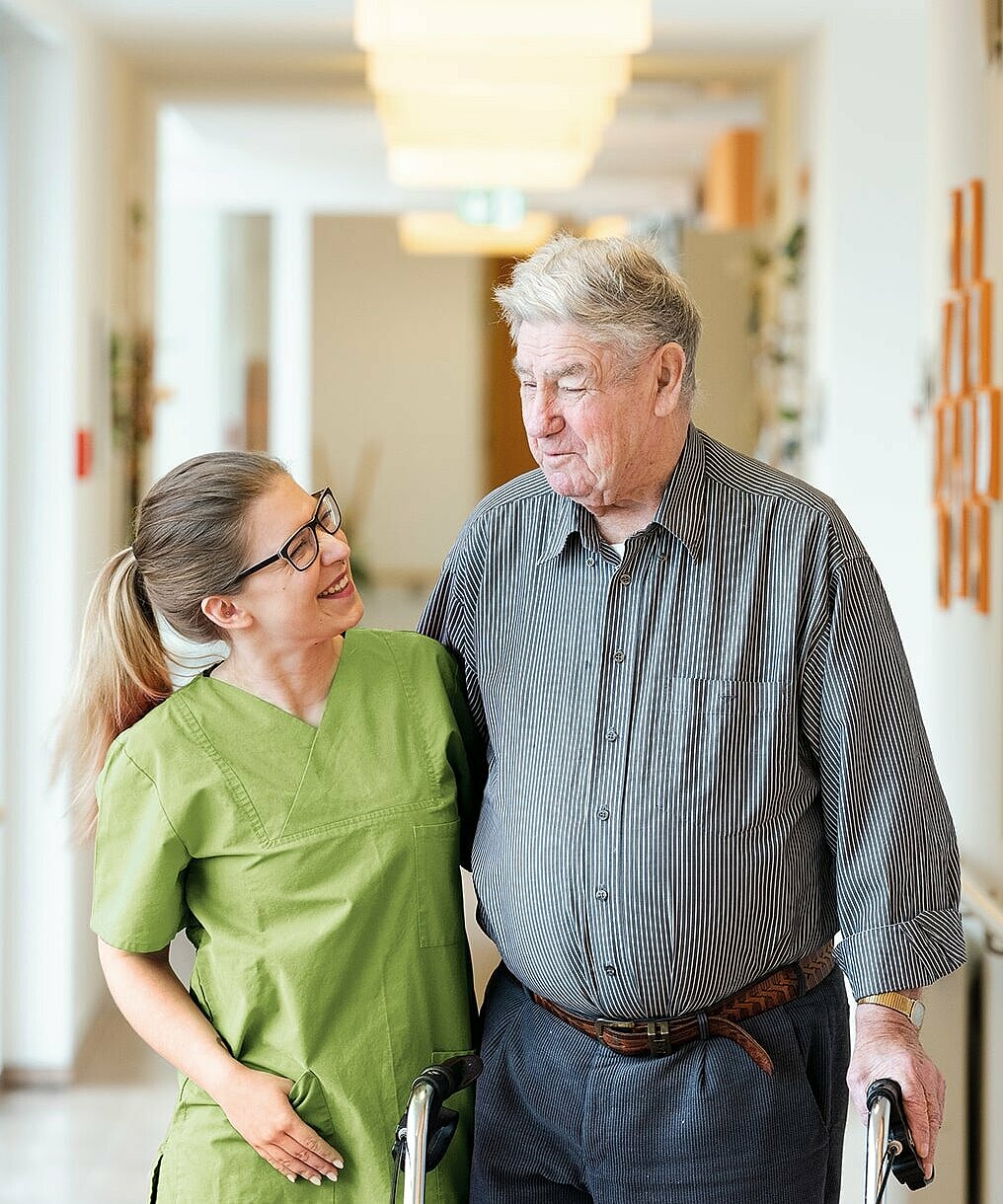 Eine lächelnde junge Frau in medizinischer Kleidung unterstützt einen älteren Mann mit Gehhilfe beim Gehen durch einen hellen Flur.